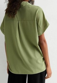Camicia verde a maniche corte con motivo a quadri, colletto, dettaglio della piega posteriore e vestibilità rilassata. Texture del tessuto liscia e visibile.