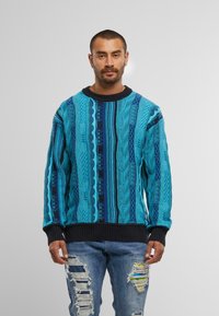 Maglione lavorato a maglia multicolore con motivi turchesi, blu e neri, caratterizzato da trecce testurizzate e polsini e orlo a costine.