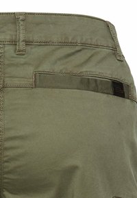 Olive green pants with a back welt pocket featuring a fabric trim and a small black brand label.