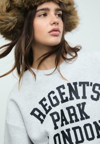 Šedá mikina s výrazným černým nápisem "REGENT'S PARK LONDON." Modelka nosí hnědou kožešinovou čepici, má vlnité vlasy a neutrální pozadí.
