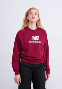 Pull bourgogne avec col rond, poignets et ourlet côtelés, présentant un logo "new balance" blanc sur le devant.