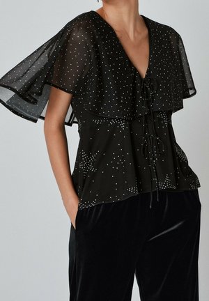 Femme portant un chemisier noir transparent à petits pois blancs, manches volantées, détails noués devant, et pantalon en velours noir, mains dans les poches.