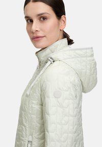 Giacca trapuntata verde chiaro con cappuccio, chiusura a zip e una patch con logo. Il tessuto ha una texture liscia con un motivo geometrico.