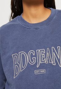 Marinblå sweatshirt med "BDG JEAN" i vita broderade bokstäver och "EST. 1998"-etikett. Mjuk textur med en klassisk rundhalsdesign.