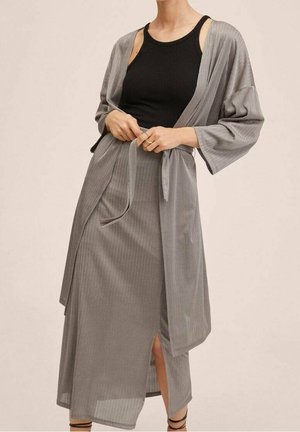 Kvinde iført en lang, grå, koksrandet nederdel og matchende kimono, bundet i taljen, over en sort ærmeløs top, stående foran en ensfarvet baggrund.