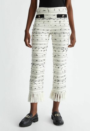 Femme portant un pantalon blanc court avec des rayures horizontales noires, des ourlets à franges, des rabats de poche noirs avec des boutons et des mocassins noirs avec détail doré.