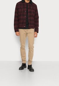 Mężczyzna ubrany w ciemnoczerwoną kratkowaną marynarkę z czarnym kołnierzem z owczej wełny, czarną koszulę, beżowe spodnie i czarne skórzane buty, stojący przy białej ścianie.