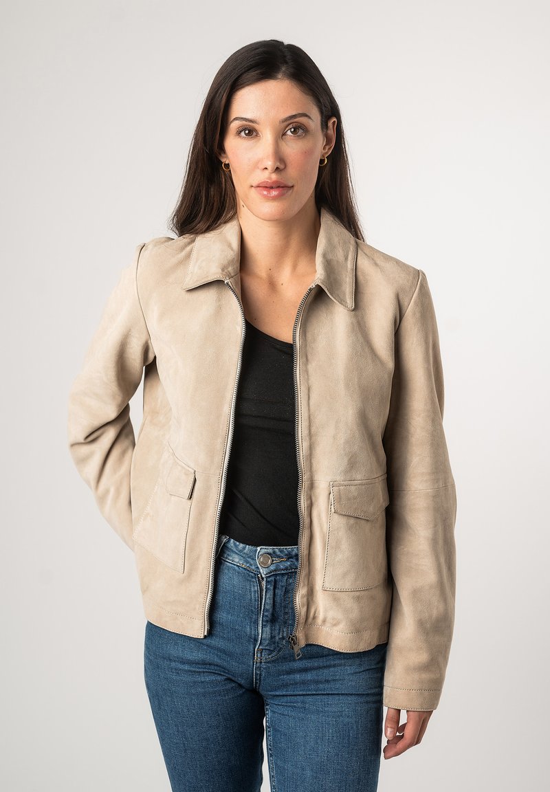 Beige Lederjacke mit kragenloser Hals, Frontreißverschluss und zwei Brusttaschen. Glatte Textur, taillierte Passform, kombiniert mit blauen Jeans.