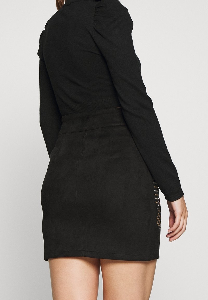 Femme portant un haut noir moulant à manches longues et une mini-jupe noire taille haute avec des découpes sur les côtés.