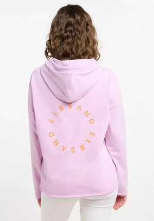 A person with curly hair wears a light pink hoodie featuring orange text in a circular design on the back, standing against a plain background.