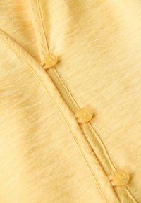 Žlutá látka s jemnou texturou a decentním leskem. Obsahuje několik žlutých knoflíků podél légu a pečlivě vyšitý šev.