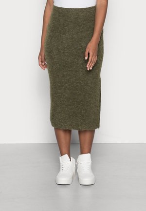 Kvinde iført en knælængde olivengrøn blød nederdel og hvide høje sneakers, der står mod en ensfarvet lys baggrund.
