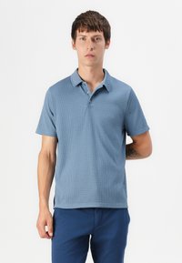 Svetlo modra polo majica s kratkimi rokavi in teksturiranim vzorcem, z gumbasto ovratnikom. Nosi se z temno modrimi hlačami.