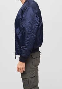 Námořnická modrá bomber bunda s lesklým saténovým povrchem, žebrovanými manžetami a postranní kapsou na zip. Nosená přes olivové cargo kalhoty.