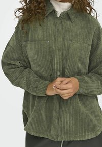 Zielona koszula z kordurowego materiału z zapięciem na guziki z przodu, dwoma kieszeniami na piersi, żeberkową teksturą i luźnym krojem. Posiada kołnierzyk i zatrzaski.