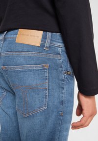 Modré džíny z denimu s kontrastním oranžovým prošíváním, koženou cedulkou na zadní straně a užším střihem. Obsahují pět kapes.