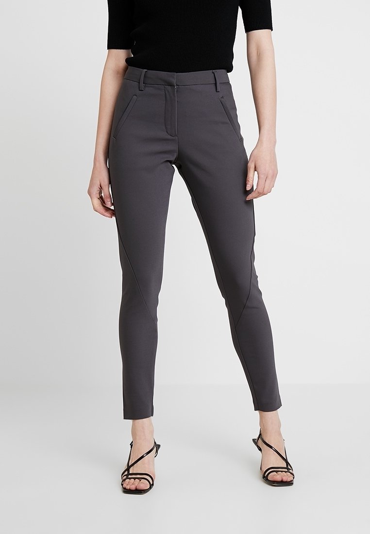 Pantalon gris ajusté en tissu extensible, présentant des coutures inclinées et des poches avant, associé à des talons noirs à lanières.