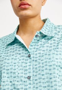 Cămașă cu guler de culoare teal deschis, având un model geometric în gri închis, cu închidere cu nasturi în față și partea interioară a gulerului alb. Textură de țesătură moale.