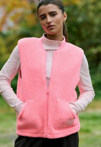 Roza telečja jopa z teksturirano, puhasto tkanino, zadrgo, rebrastim ovratnikom in dvema sprednjima žepoma. Nošena čez dolg rokav.