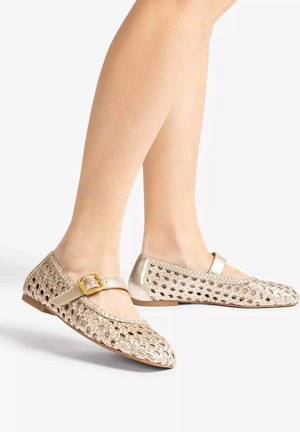 Feet wearing gold woven Mary Jane flats with a gold buckle on a white background.