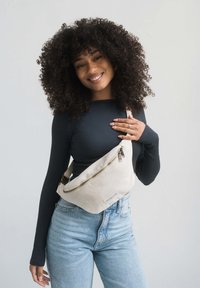 Beige fabric fanny pack with a gold zipper, worn across the torso. Features clean lines, a small logo patch, and a simple strap.