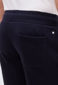 Κοντινό πλάνο σε μπλε παντελόνι ναυτικού με ελαστική μέση και τσέπη πίσω, φορεμένο με λευκό πουκάμισο μέσα.