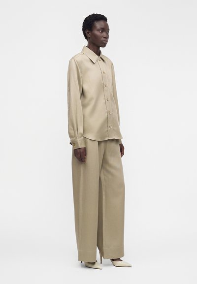 Μοντέλο φοράει μπεζ πουκάμισο oversized με κουμπιά και αντίστοιχες φαρδιές παντελόνες με μπεζ μυτερές ψηλοτάκουνες γόβες, στέκεται μπροστά σε απλό λευκό φόντο.