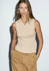 Woman with long hair wears a beige sleeveless V-neck top and loose tan pants, hands in pockets, standing against a plain light background.