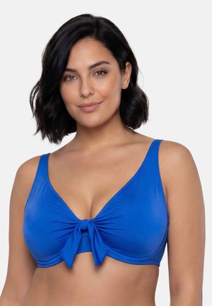 Kvinde med skulderlangt sort hår iført en blå bikini top med bindebånd foran, smilende mod en ensfarvet hvid baggrund.