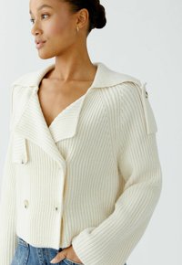 Frau trägt eine cremefarbene, grob gestrickte Cardigan mit breiten Revers und Knopfdetails, kombiniert mit blauen Jeans, die Hände in den Taschen.