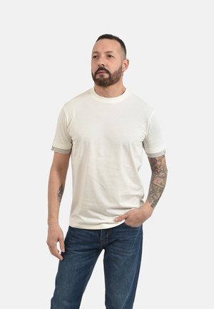 Uomo con capelli corti e barba, in piedi con la mano in tasca, indossa una maglietta bianca e jeans blu, mostrando un tatuaggio colorato sulla manica del braccio destro.