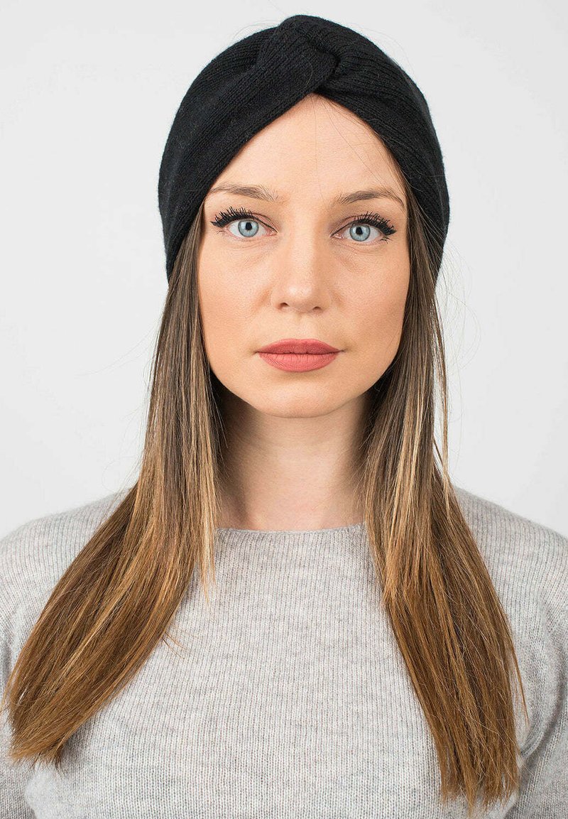 Černý pletený turbanový čelenka s atypickým designem vpředu, nošený přes dlouhé hnědé vlasy. Na obrázku je vidět světle šedý svetr.