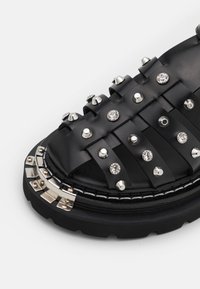 Sandale en cuir noir avec clous argentés et strass transparents sur plusieurs brides et une semelle épaisse avec ornements métalliques.