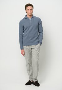 Blauer gerippter Strickpullover mit einem Viertel-Reißverschluss, kombiniert mit hellgrauen, maßgeschneiderten Hosen und dunkelbraunen Wildleder-Schlupfschuhen. Schlichte, klare Linien über das gesamte Outfit.