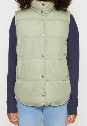 Chaleco acolchado verde claro con botones de presión, usado sobre una camisa de manga larga azul marino y vaqueros azul claro, mostrado desde los hombros hasta las caderas.