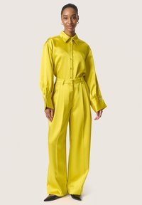 Set in satin giallo composto da camicia e pantaloni a gamba larga, caratterizzato da un colletto, dettagli con bottoni e maniche ampie con accenti sui polsini. Scarpe nere a punta.