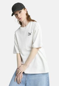 White cotton t-shirt with short sleeves and a black Puma logo on the left chest, paired with a black cap and a light blue denim skirt.