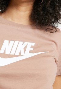 Hnědý bavlněný tričko s velkým bílým logem "NIKE" na hrudi a zakřiveným bílým šipkovým motivem pod ním. Krátké rukávy, kulatý výstřih.