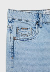 Jeans de mezclilla azul claro con un bolsillo frontal, costuras en contraste y un pequeño acento de logotipo metálico en el bolsillo. Textura suave, diseño casual.