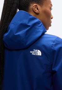 Veste imperméable bleue avec capuche. Présente un logo blanc proéminent dans le dos, une texture lisse et une coupe ajustée.