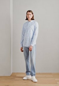Camicia button-up azzurro chiaro con un leggero motivo a nuvole, abbinata a jeans in denim chiaro e sneakers bianche, in piedi su un pavimento di legno.