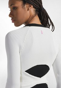 Weißes, geripptes Langarmshirt mit ausgeschnittenem Rücken und schwarzen Akzenten. Es hat eine figurbetonte Silhouette und ein pinkfarbenes Etikett nahe dem Halsausschnitt.