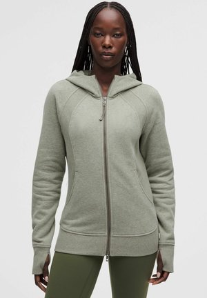 Frau trägt ein hellgraues Kapuzensweatshirt mit Reißverschluss und Daumenlöchern sowie olivgrüne Leggings und steht vor einem schlichten grauen Hintergrund.
