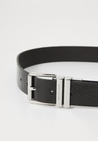 Musta nahkavyö, jossa on sileä pinta ja hopeanvärinen metallinen solki, jossa on kaiverrettu brändäys. Suorakulmainen muoto, yksinkertainen lenkki ja reiät koon säätämiseksi.