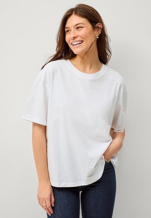 T-shirt in cotone bianco con maniche corte, scollatura rotonda, vestibilità comoda e texture morbida; indossato con jeans di denim scuro. Nessun motivo o dettaglio.