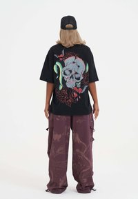 Sort oversized t-shirt med et kraniumgrafik og blomstermotiv på ryggen, parret med løse lilla cargo-bukser med subtile mønstre.
