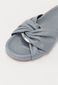 Sandale à glissière en cuir bleu avec un design noué et un accent circulaire en argent. Texture lisse et forme de bout arrondi.