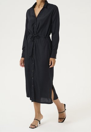 Kvinde iført en langærmet, navy blå button-down midi-kjole med taljebånd og sideslidser, kombineret med sorte hælede sandaler.