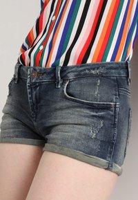 Tummansiniset farkkushortsit haalistuneella pesulla, joissa on käännetyt reunat, kulutetut yksityiskohdat, etutaskut ja nappikiinnitys.