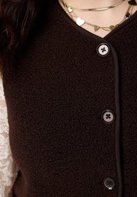 Bruine fleecevest met een V-hals en vijf knopen aan de voorkant. Voorzien van een gestructureerde stof en een decoratieve laagjeskantmouw.